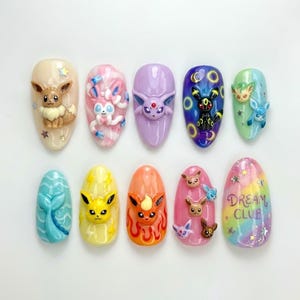 May include: A set of ten press-on nails featuring 3D character embellishments inspired by Eevee and its evolutions. The designs include various colors, patterns, and textures, with one nail displaying the text DREAM CLUB in purple lettering.
