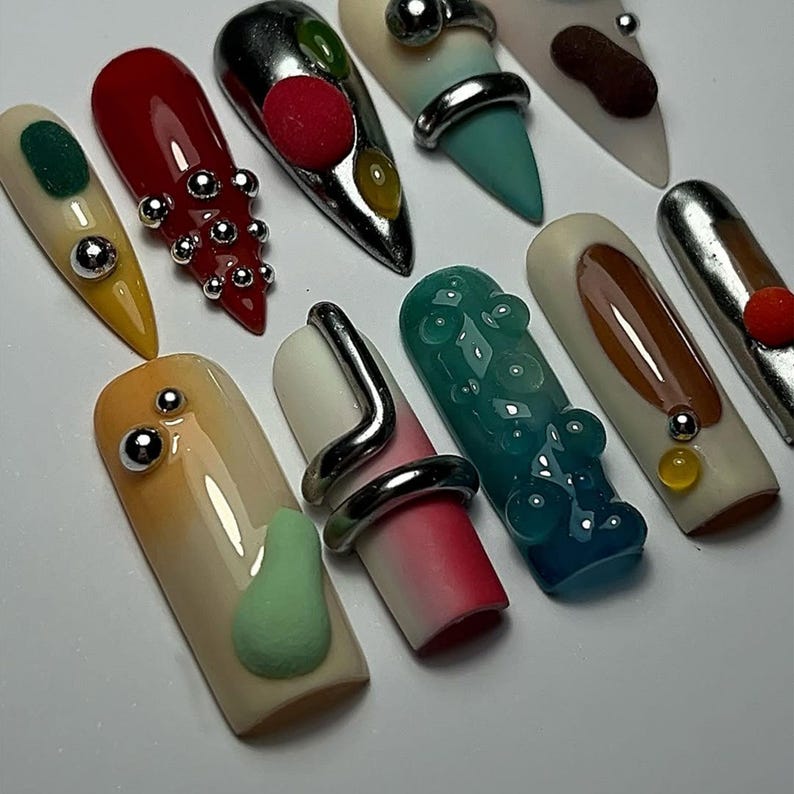 May include: A collection of artificial nails with various designs. Some nails are painted in solid colors like red, yellow, and teal, while others feature abstract designs, metallic accents, and 3D elements. The nails are arranged on a white surface.