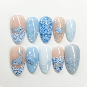 May include: A set of ten artificial nails with a marine theme. The nails feature various designs, including whales, waves, and decorative patterns in shades of blue, white, and beige. Some nails have 3D whale designs.