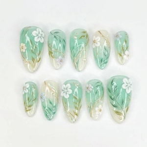 May include: A set of ten artificial nails with a floral design. Each nail is painted with a light green base, white flowers, and gold accents. The nails are almond-shaped and feature a gradient effect.