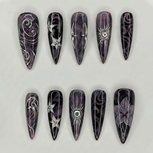 May include: A set of ten stiletto-shaped press-on nails featuring a deep purple and black marbled base. The designs include raised silver metallic accents, intricate swirling patterns, star-like floral motifs, vertical lines, and a central sunburst.