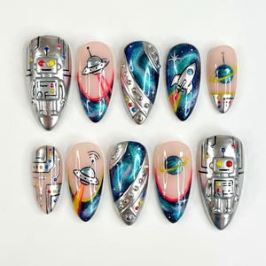 May include: A set of ten artificial nails with an outer space theme. Designs include a silver robot, a UFO, a rocket ship, and a planet. The nails feature a variety of colors, including silver, blue, pink, and yellow. The nails are almond-shaped.