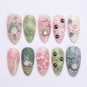 Peut inclure: Un ensemble de dix faux ongles en forme d'amande avec des motifs en relief 3D. Les illustrations incluent des personnages Totoro gris, des fleurs de cerisier roses, des feuilles vertes, des suies noires et des gouttes d'eau sur fond pastel.
