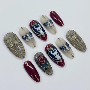 May include: A collection of oval-shaped press-on nails with intricate designs. The nails feature a variety of colors, including burgundy, gray, and cream, with gold accents. Some nails showcase a white bull design, a blue butterfly, and a red rose.