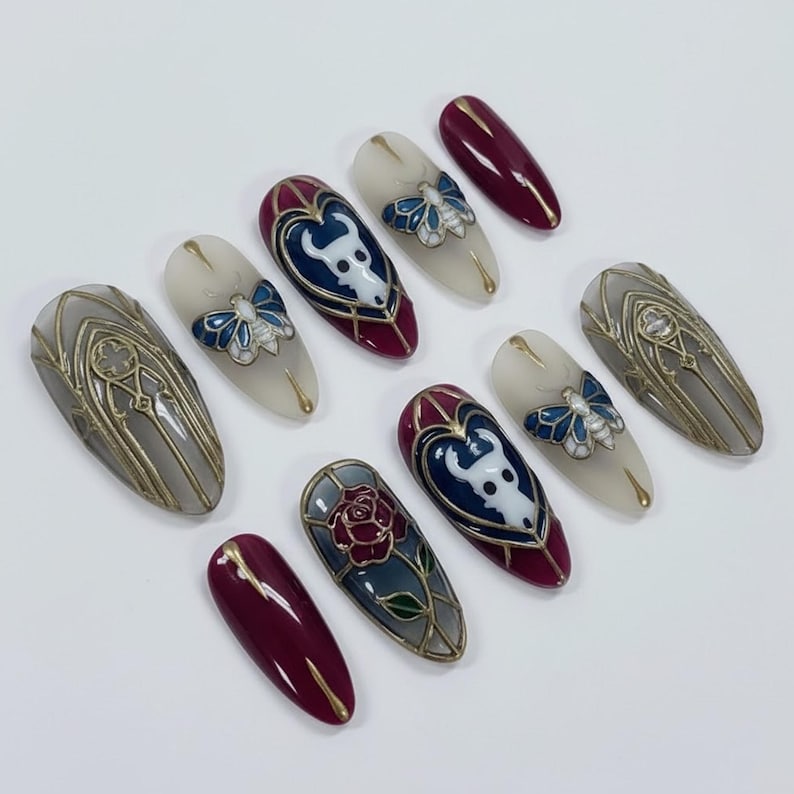 May include: A collection of oval-shaped press-on nails with intricate designs. The nails feature a variety of colors, including burgundy, gray, and cream, with gold accents. Some nails showcase a white bull design, a blue butterfly, and a red rose.