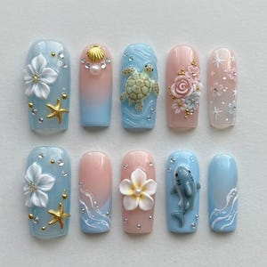 May include: A set of ten press-on nails featuring ocean-themed designs including 3D white flowers, gold starfish, a sea turtle, a shark, pink roses, and white wave patterns. The nails are decorated with small rhinestones, gold beads, and pearls.