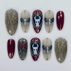 May include: A set of ten artificial nails with various designs. Some nails feature a gray gothic window design with gold accents. Others showcase a white character, a blue butterfly, and a red rose. The nails are oval-shaped and have a glossy finish.