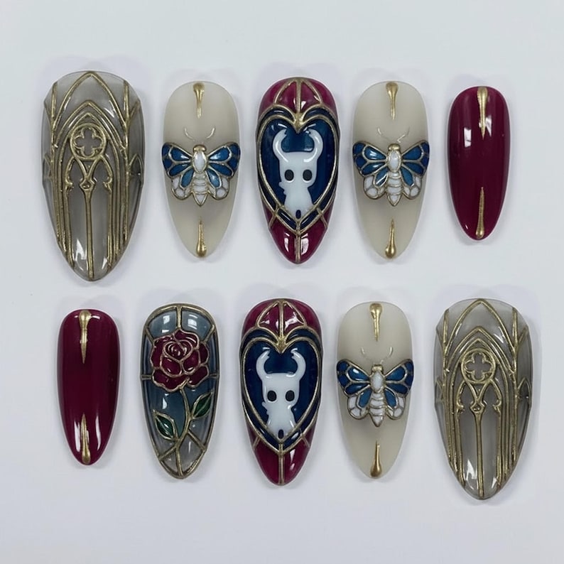 May include: A set of ten artificial nails with various designs. Some nails feature a gray gothic window design with gold accents. Others showcase a white character, a blue butterfly, and a red rose. The nails are oval-shaped and have a glossy finish.