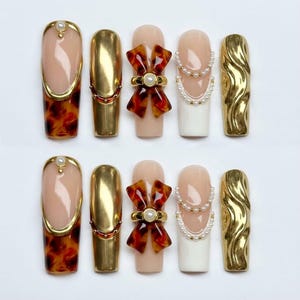 Peut inclure: Un ensemble de dix faux ongles aux designs variés. Les ongles présentent des couleurs or, écaille de tortue, nude et blanc. Certains ongles sont ornés de perles, de nœuds et d'un motif doré ondulé.