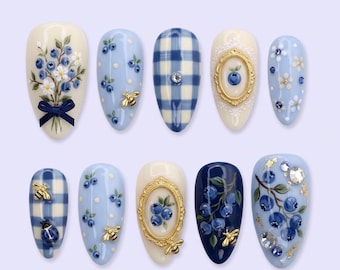 May include: A collection of ten almond-shaped artificial nails with various designs. The designs include blueberries, bees, floral patterns, and a gingham pattern. The color palette consists of blue, white, and gold accents. Some nails feature embellishments like rhinestones and gold frames.