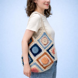 Könnte beinhalten: Eine handgefertigte Häkeltasche mit Patchwork-Design. Die Tasche hat Quadrate in Blau-, Orange-, Rosa- und Cremetönen mit einem cremefarbenen Riemen. Die Tasche wird über der Schulter getragen.