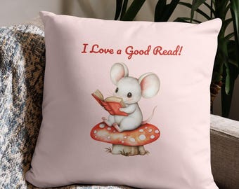 Może przedstawiać: Jasnoróżowa kwadratowa poduszka z napisem "I Love a Good Read!". Zawiera akwarelową ilustrację białej myszy czytającej książkę, siedzącej na czerwono-białym grzybie. Poduszka leży na szarej kanapie.