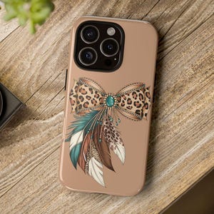 Op de afbeelding: Een beige telefoonhoesje met een luipaardprint strik, een turquoise edelsteen en bungelende kralen. Onder de strik bevinden zich blauwe, bruine en witte veren. Op een houten oppervlak.