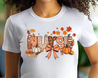 May include: A white short-sleeved t-shirt featuring a graphic print of the word NURSE in large, patterned letters. The design includes autumn-themed elements such as orange maple leaves, two pumpkins, a large peach-colored bow, and small flowers.