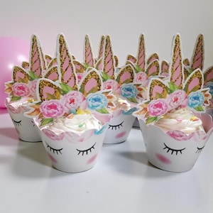 Op de afbeelding: Een verzameling cupcakes met eenhoornthema met witte glazuur, bloemdecoraties en papieren toppers. De cupcakes zitten in witte papieren cups met gesloten oogontwerpen. Twee ballonnen, één blauw en één roze, staan op de achtergrond.