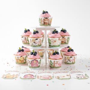 Può includere: Un espositore bianco a più livelli presenta cupcakes con glassa rosa e mirtilli. I pirottini dei cupcake presentano disegni di animali della fattoria. Carte a tema fattoria abbinate sono esposte davanti.