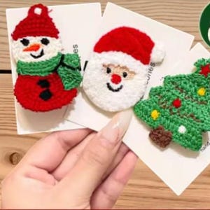 Könnte beinhalten: Gehäkelte Haarspangen mit Weihnachtsmotiven. Das Set enthält einen Schneemann mit roter Mütze und grünem Schal, ein Gesicht des Weihnachtsmanns mit weißem Bart und roter Mütze sowie einen grünen Weihnachtsbaum mit roten und gelben Ornamenten. Jede Spange ist auf einer weißen Karte.