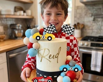 May include: A white frosted cake decorated with race car toppers, checkered flags, and colorful balloons. The cake has a black and white racing stripe around the base and the name "Reid" in gold script. A child is holding the cake.