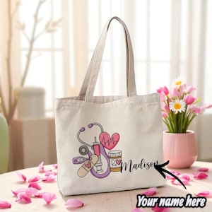Pode incluir: Saco tote creme com um gráfico médico, incluindo um estetoscópio, coração, seringa e frasco de medicamentos. O saco é personalizado com um nome e é adequado para profissionais ou estudantes de medicina.