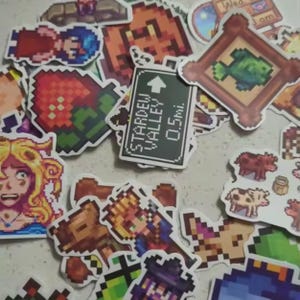 Peut inclure: Un assortiment d'autocollants color&eacute;s en pixel art repr&eacute;sentant des personnages, des objets et des sc&egrave;nes du jeu vid&eacute;o Stardew Valley. Comprend un panneau indiquant "Stardew Valley 0.5 mi.", une fraise et un poisson encadr&eacute;.