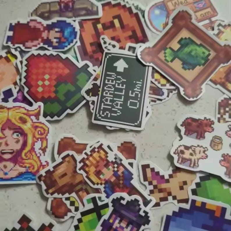 Peut inclure: Un assortiment d'autocollants color&eacute;s en pixel art repr&eacute;sentant des personnages, des objets et des sc&egrave;nes du jeu vid&eacute;o Stardew Valley. Comprend un panneau indiquant "Stardew Valley 0.5 mi.", une fraise et un poisson encadr&eacute;.