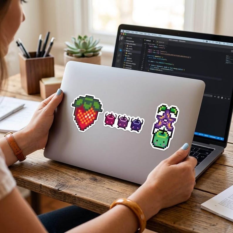 Peut inclure: Un ordinateur portable orn&eacute; d'autocollants en pixel art. Les autocollants repr&eacute;sentent une fraise rouge et verte, trois petites cr&eacute;atures violettes et une plante verte et violette. L'ordinateur portable est ouvert sur un bureau en bois.