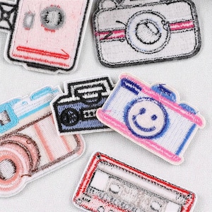 Peut inclure: Plusieurs écussons brodés représentant des appareils photo. Les écussons présentent différents modèles d'appareils photo dans des tons de rose, de bleu et de noir, avec des détails tels que des objectifs et un visage souriant. Les écussons sont bordés de noir.
