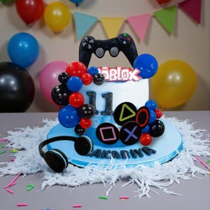 May include: A birthday cake decorated with a black video game controller, the word "ROBLOX", and the number "11". The cake is adorned with red, blue, and black spherical decorations, and a pair of black headphones. The cake is on a blue base.