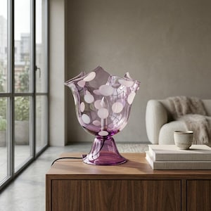 May include: A translucent purple glass table lamp with a wide, ruffled rim and a pattern of opaque white circular spots. The lamp features a central light bulb socket and a flared pedestal base, resting on a flat surface with a black power cord.