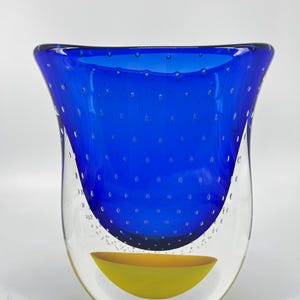 May include: A contemporary art glass vase featuring a striking blue interior with air bubbles. The exterior is clear, with a yellow bowl-shaped element at the base. The vase has a distinctive, curved form.
