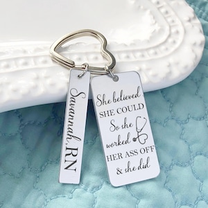 May include: A silver keychain with a heart-shaped ring and two rectangular tags. One tag reads "Savannah, RN" in script. The other tag has the text "She believed SHE COULD So she worked HER ASS OFF & she did" with a stethoscope graphic.