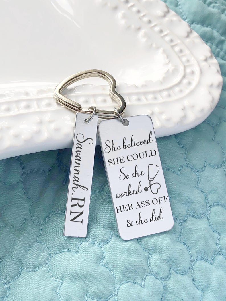 May include: A silver keychain with a heart-shaped ring and two rectangular tags. One tag reads "Savannah, RN" in script. The other tag has the text "She believed SHE COULD So she worked HER ASS OFF & she did" with a stethoscope graphic.