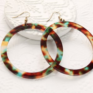 May include: A pair of large, round hoop earrings with a tortoiseshell pattern. The earrings feature a mix of brown, orange, and teal colors. The hooks are gold-colored.