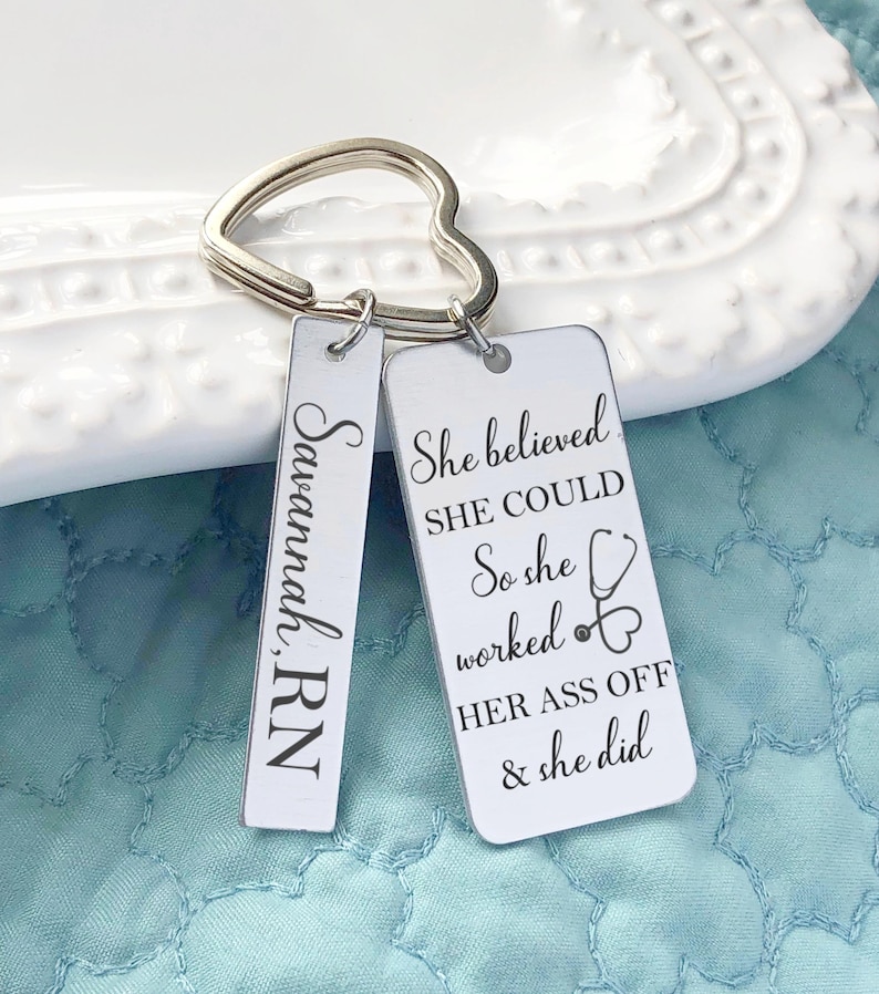 May include: A silver keychain with a heart-shaped ring. It features two rectangular tags. One tag reads "Savannah, RN" vertically. The other tag has the text "She believed she could, So she worked HER ASS OFF & she did."