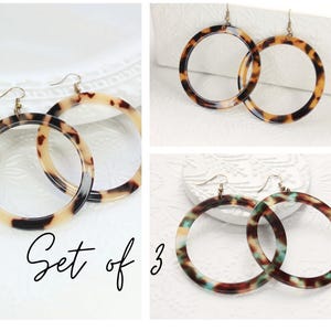 May include: Set of 3 pairs of large hoop earrings. The earrings feature a tortoiseshell pattern in shades of brown, tan, and black. One pair has a brown and tan pattern, another has a brown and black pattern, and the third pair has a brown, teal, and tan pattern.
