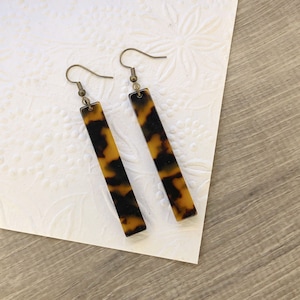 May include: A pair of long, rectangular earrings with a tortoiseshell pattern. The earrings have a gold-tone finish and are made of a lightweight material.