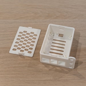 May include: A white plastic rectangular enclosure with a separate perforated cover. The enclosure has a series of horizontal slots and a curved side with three holes. The cover has a honeycomb pattern of small holes.