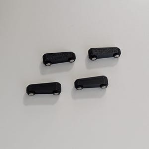 May include: Four small, black rectangular objects with rounded edges and two small, silver-colored circular accents on the bottom. The objects are evenly spaced against a white background. The items appear to be small magnets.