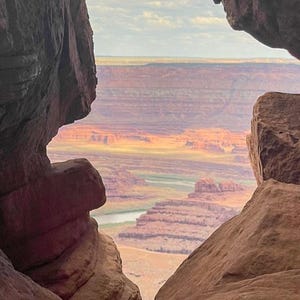 Könnte beinhalten: Eine malerische Aussicht, eingerahmt von rötlich-braunen Felsformationen. Die Landschaft zeigt einen Canyon mit Schichten aus rotem und orangefarbenem Gestein, einen Fluss und einen bewölkten Himmel. Der natürliche Bogen schafft eine einzigartige Perspektive.