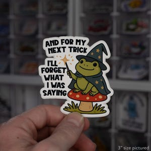 May include: A sticker featuring a cartoon frog dressed as a wizard, holding a wand and sitting on a mushroom. The sticker has the text "AND FOR MY NEXT TRICK I'LL FORGET WHAT I WAS SAYING". The sticker is approximately 3 inches in size.