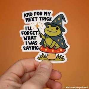 May include: A die-cut sticker featuring a green frog wearing a dark blue wizard hat and cape with yellow stars, sitting on a red mushroom with white spots. The frog holds a magic wand. Text reads: AND FOR MY NEXT TRICK I'LL FORGET WHAT I WAS SAYING.