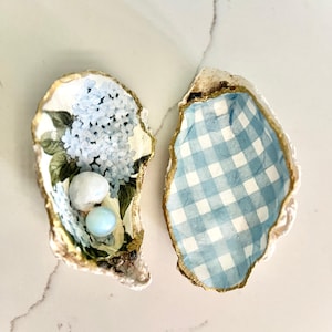 May include: Two decorative oyster shells. One shell features a blue hydrangea design with two small, round, light-coloured objects. The other shell has a blue and white gingham pattern. Both shells have gold-painted edges.