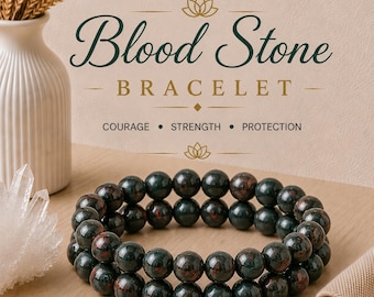 Puede incluir: Una pulsera de doble hilo hecha de cuentas de piedra de sangre verde oscuro y rojo. El texto sobre la pulsera dice Blood Stone BRACELET, COURAGE, STRENGTH, PROTECTION. La pulsera está colocada cerca de un jarrón blanco y un cristal.