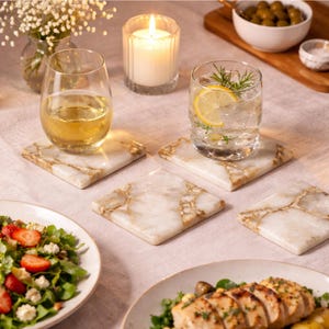 May include: A set of four square marble coasters featuring a white base with prominent gold and brown veining. The coasters have a smooth, polished finish and are arranged on a light-colored fabric surface, supporting two clear glass tumblers.