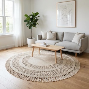 May include: A round, beige macrame rug with a fringed edge sits on a hardwood floor. The rug is in a living room setting with a light-colored sofa, wooden coffee table, and a potted plant.