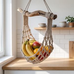 May include: A macrame fruit hammock suspended from a natural wood branch. The beige woven basket is filled with ripe bananas, mangoes, and oranges. The kitchen setting features a wooden countertop, white tile backsplash, and a window.