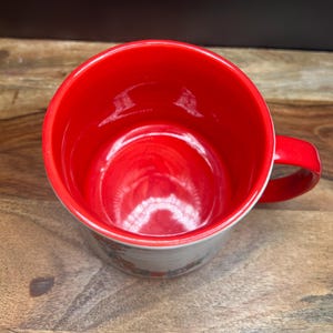 May include: A ceramic mug with a bright red interior and handle. The exterior features a floral design around the base. The mug is set against a wooden surface.