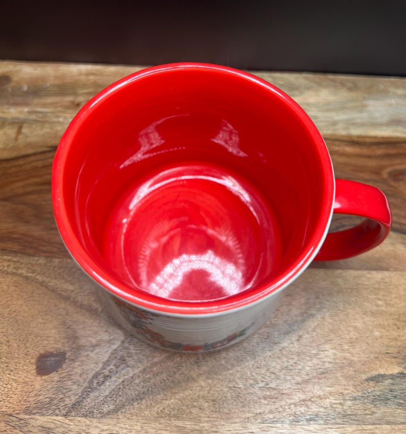May include: A ceramic mug with a bright red interior and handle. The exterior features a floral design around the base. The mug is set against a wooden surface.