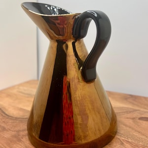 May include: A gold-colored ceramic pitcher with a black handle and interior. The pitcher has a glossy finish and a wide spout. The handle is curved and attached to the side of the pitcher. The pitcher is sitting on a wooden surface.