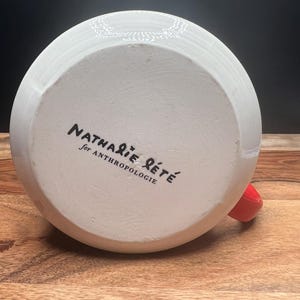 May include: White ceramic mug with a red handle. The bottom of the mug features the text "Nathalie Lete for Anthropologie" in black script. The mug is set on a wooden surface.